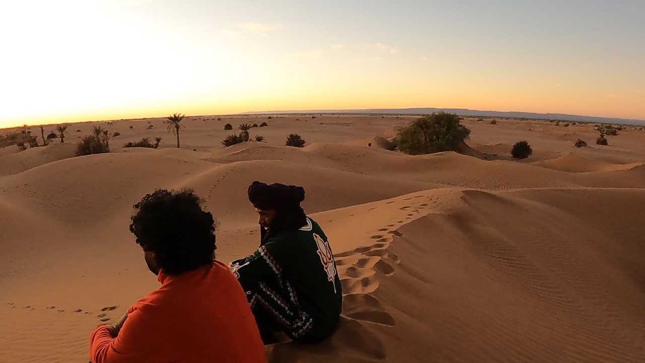 Two people seated on sand dunes at sunset