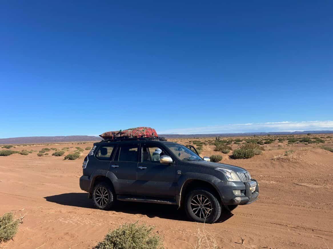 Four-wheel-drive vehicle carrying luggage across a sandy desert track under a blue sky