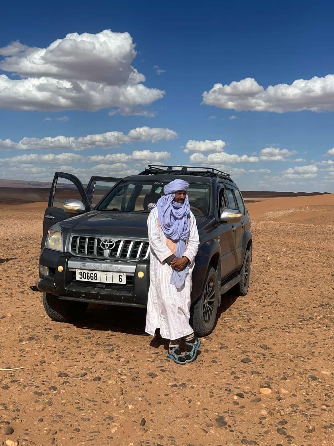 Man in traditional desert clothing standing beside a four-wheel-drive vehicle in a rocky desert landscape