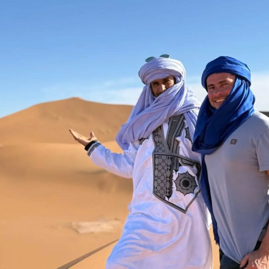 Two men wearing desert headscarves standing in front of golden sand dunes